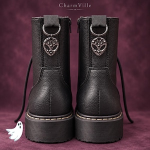 May include: A pair of black textured boots with a side zipper and a black fabric pull tab featuring a metal D-ring. An alien head charm with dark studs hangs from each D-ring. The text CharmVille is visible at the top center.