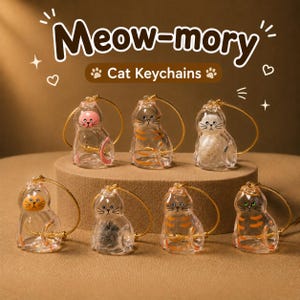 May include: Seven clear glass cat-shaped keychains with gold-toned wire loops. Each keychain features a unique cat face and internal detail, including orange stripes, pink accents, and textured fillings. The text Meow-mory Cat Keychains is visible.