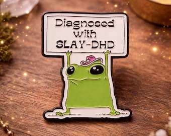 Puede incluir: Un pin de rana verde con un sombrero de vaquero rosa que sostiene un letrero que dice "Diagnosed with SLAY-DHD". El pin tiene un borde blanco y está sobre un fondo de madera.