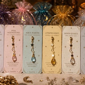 May include: Six charm necklaces with gold-colored chains, black stars, and crescent moons, each with a different gemstone. The cards behind the charms have text: "VIOLET AURA", "PINK MOONSTONE", "AQUA MARINE", "CITRINE LIGHT", "MOON QUARTZ", and "MOON MIST".