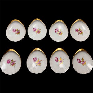 May include: Eight white porcelain shell-shaped dishes with gold rims. Each dish features a pink rose and gold leaf design. The dishes are arranged on a black background, showcasing their elegant details.