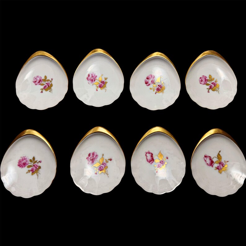 May include: Eight white porcelain shell-shaped dishes with gold rims. Each dish features a pink rose and gold leaf design. The dishes are arranged on a black background, showcasing their elegant details.