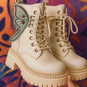 May include: Cream-colored lace-up boots with a butterfly wing accent. The boots have a chunky sole with a pearl trim and gold eyelets. The butterfly wing is green with red accents.