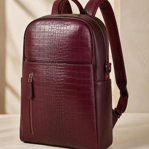 May include: A deep burgundy backpack featuring a textured crocodile-embossed pattern on the front panel. The bag includes a vertical zippered front pocket with a metallic pull, a main zippered compartment, a top handle, and adjustable shoulder straps.