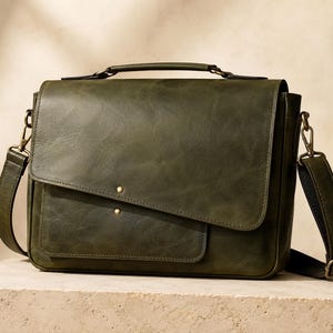 Puede incluir: Un bolso tipo mensajero de cuero verde oliva con asa superior, correa de hombro ajustable con herrajes de metal en tono latón y un cierre de solapa frontal asegurado con dos pequeños remaches de latón. Tiene una forma rectangular.