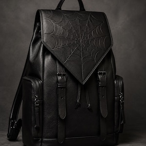 May include: A black backpack with a textured finish featuring a prominent embossed spiderweb design on the triangular top flap. The bag includes two vertical buckled straps, a drawstring closure, and two side pockets with zipper hardware.