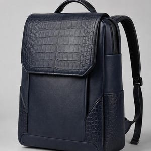 Puede incluir: Una mochila de color azul marino oscuro con una silueta rectangular estructurada y cierre de solapa. El exterior presenta una combinación de texturas lisas y estampadas de cocodrilo, un asa superior y correas ajustables para los hombros.