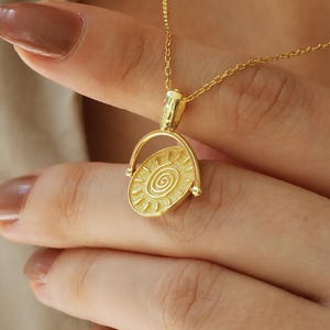 May include: A gold-colored pendant necklace featuring a circular pendant with a spiral design. The pendant is suspended from a delicate chain. The necklace is displayed against a neutral background.