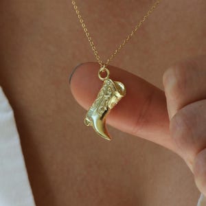 Peut inclure: Un collier pendentif doré avec un charme en forme de botte de cowboy. Le charme est suspendu à une fine chaîne dorée. La botte présente des motifs en relief détaillés et un bout pointu. Le collier est présenté sur une chemise blanche.