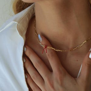 Op de afbeelding: Een goudkleurige ketting met een rechthoekig schakelontwerp. Een klein rood touwtje is aan de ketting vastgemaakt. De ketting wordt gedragen over een wit overhemd.