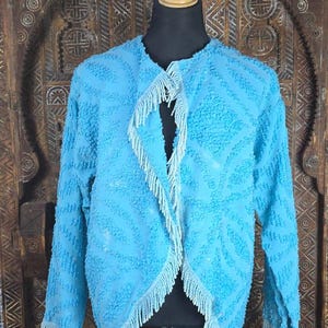 May include: A bright turquoise jacket with a textured, raised pattern across the fabric. The garment features long sleeves and an open front design, finished with light-colored fringe trim along the edges of the collar, front opening, and cuffs.