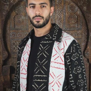 Könnte beinhalten: Eine schwarze Langarmjacke mit weißen vertikalen Bahnen auf der Vorderseite, die mit roten geometrischen Stickmustern versehen sind. Die Jacke hat einen Kragen und einen schwarzen Grundstoff, der mit weißen, sich wiederholenden Symbolen verziert ist.