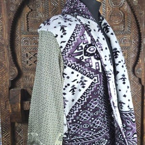 May include: A long-sleeved wrap garment featuring a white and deep purple geometric pattern on the main body and a contrasting light olive green sleeve with a fine black repeating print. The hem is finished with long white fringe detailing.