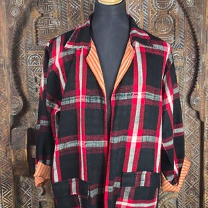 Puede incluir: Una chaqueta de manga larga con un patrón de cuadros en negro, rojo y blanco. La prenda tiene un cuello con muescas, dos bolsillos delanteros y un forro a rayas naranjas y blancas visible en los puños y solapas. El dobladillo tiene flecos.