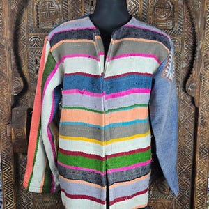 Puede incluir: Un abrigo de manga larga con un vibrante patrón de rayas horizontales en tonos rosa, azul, verde, amarillo, naranja y gris. La prenda tiene un escote sin cuello, un diseño frontal abierto y un pequeño detalle bordado en la manga izquierda.