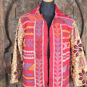 Puede incluir: Una chaqueta de manga larga con una base de color rojo vibrante, adornada con intrincados bordados geométricos y abalorios multicolores. Las mangas presentan un patrón floral en contraste de color amarillo y granate, mientras que los paneles frontales están decorados con filas verticales de triángulos y bandas horizontales en azul, amarillo y blanco.