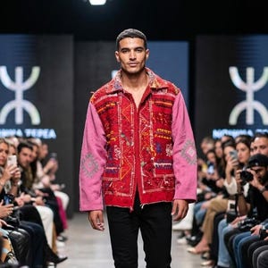 Könnte beinhalten: Eine langärmelige Jacke mit einer leuchtend roten Vorderseite, die mit komplexen, mehrfarbigen geometrischen Stickereien verziert ist, sowie einfarbigen rosa Ärmeln. Die Jacke hat einen Kragen, einen Frontreißverschluss und eine schmale schwarze Hose.