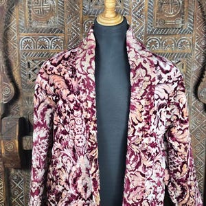 Könnte beinhalten: Ein langärmeliger Blazer mit einem floralen Damastmuster in tiefem Burgunderrot und Creme. Das Kleidungsstück verfügt über einen Schalkragen, ein offenes Vorderteil und eine strukturierte, samtartige Oberfläche, präsentiert auf einer Schneiderpuppe.