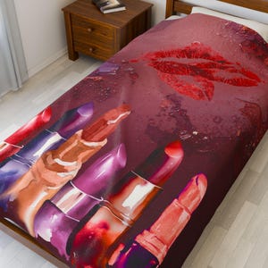 Op de afbeelding: Een bordeauxkleurig dekbedovertrek met een aquarelontwerp van rode lippenstiften en een grote rode lipafdruk. De lippenstiften zijn in verschillende tinten rood, roze en paars. Het beddengoed ligt op een houten bedframe.