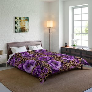 May include: A bed comforter featuring a vibrant purple floral pattern layered over a brown and black leopard print background. The bedding is draped over a bed frame with two white pillows positioned at the head of the mattress.