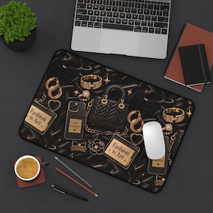 May include: A rectangular desk mat featuring a black satin background with a gold-toned pattern of handbags, jewelry, and smartphones. The design includes text reading Fashion is Art and Style Inspiration, with a white computer mouse placed on top.