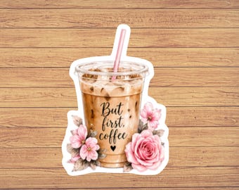 May include: A clear plastic cup filled with iced coffee, featuring a pink straw and the black text But first, coffee with a small heart. The cup is decorated with light pink floral illustrations of roses and leaves on the bottom sides.