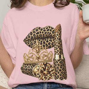 May include: A light pink t-shirt featuring a graphic print of lips, a lipstick tube, and a tiara, all decorated with a brown and black leopard print pattern. The design also includes a large, faceted golden gemstone accent at the base.