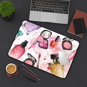 May include: A rectangular mousepad featuring a watercolor design of makeup items. The design includes a perfume bottle, blush compacts, a makeup brush, nail polish, and lipstick, all in shades of pink and purple, with a white background.