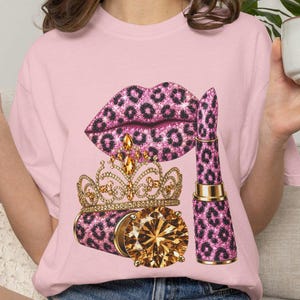 Pode incluir: Uma t-shirt rosa claro com uma estampa gráfica de lábios com padrão de leopardo rosa e preto, uma coroa dourada com gemas cor de âmbar e um batom com o mesmo padrão de leopardo e uma gema grande cor de âmbar na base.