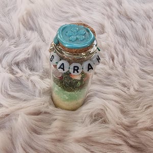 Könnte beinhalten: Ein kleines Glas mit einem Korken und einem türkisfarbenen Wachssiegel mit einem Blumendesign. Das Glas enthält Schichten aus Sand, grünem Moos und kleinen Steinen. Der Name "SARAH" ist mit weißen Buchstabenperlen um das Glas geschrieben.