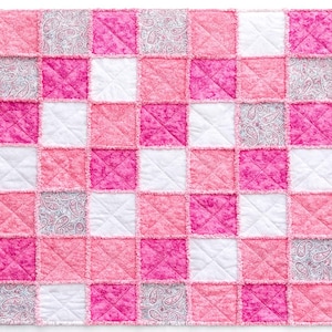 May include: A patchwork quilt in a grid pattern, featuring squares of varying pink shades, white, and paisley print. The quilt has a soft, textured appearance with visible stitching and frayed edges, creating a cozy, handmade aesthetic.