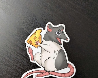Op de afbeelding: Een cartoonsticker van een rat die een stuk kaas vasthoudt. De rat is wit met zwarte en grijze markeringen, een roze staart en poten, en een verraste uitdrukking. De kaas is geel met gaten.