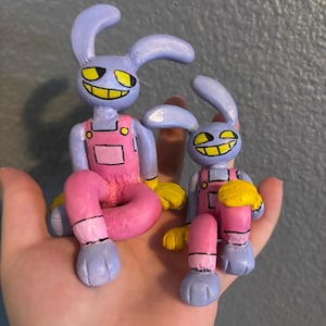 May include: Two hand-sculpted figures of purple rabbit-like characters with long ears, yellow eyes, and wide grins. Each figure wears pink overalls with yellow buttons and yellow gloves. The figures are posed in a seated position with bent legs.