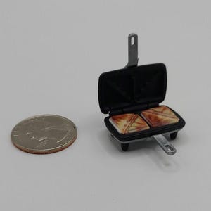 May include: Miniature black sandwich maker with two grilled sandwiches inside. The small appliance has a silver handle and is next to a coin.
