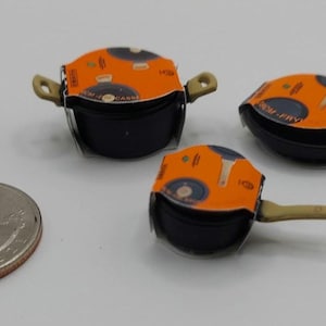 Puede incluir: Mini ollas y sartenes negras con tapas naranjas y asas color canela. Una moneda está al lado de los utensilios de cocina para comparar el tamaño. Las sartenes tienen una forma pequeña y redonda, probablemente para una casa de muñecas.