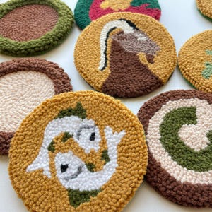 Può includere: Set di 10 sottobicchieri rotondi all'uncinetto colorati con vari disegni, tra cui un pesce, un cervo, un vortice e una lettera C. I sottobicchieri sono realizzati con filato marrone, verde, bianco e giallo.