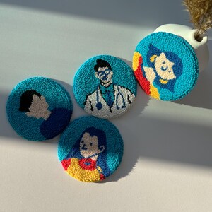 May include: Four round, turquoise coasters with embroidered illustrations. One depicts a doctor with a stethoscope, another a person with a side profile, and two others feature women. The coasters are made of a textured material.