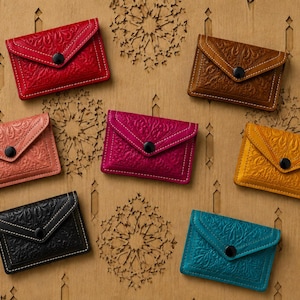 Può includere: Sette portacarte rettangolari nei colori rosso, marrone, rosa chiaro, magenta, giallo, nero e verde acqua. Ogni modello presenta una patta a busta con chiusura a bottone automatico nero e un intricato motivo floreale in rilievo sul davanti.