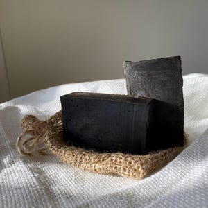 Può includere: Due saponette rettangolari marrone scuro poggiano su un piccolo sacco di juta, su uno sfondo bianco strutturato. I saponi hanno un aspetto ruvido e naturale. Il sacco di juta è fatto di fibre intrecciate di colore beige. L'illuminazione crea ombre, evidenziando le trame.