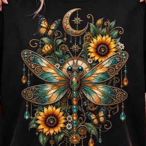May include: A black t-shirt featuring a central dragonfly graphic with teal and gold wings, surrounded by sunflowers, butterflies, a crescent moon, clock gears, and hanging crystal-like ornaments in a detailed, ornate, and colourful illustration.