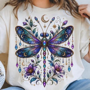 May include: A cream-colored t-shirt featuring a large, detailed graphic of a dragonfly with iridescent blue and purple wings. The design includes hanging crystal-like ornaments, crescent moons, stars, and purple flowers surrounding the insect.