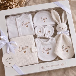 Peut inclure: Un coffret cadeau pour bébé blanc comprenant un body, un bonnet, des moufles, un bavoir et un doudou lapin, tous brodés du prénom Essia et d'un petit cœur. L'ensemble inclut un carnet texturé et une étiquette marquée AYRA CRÉATION.