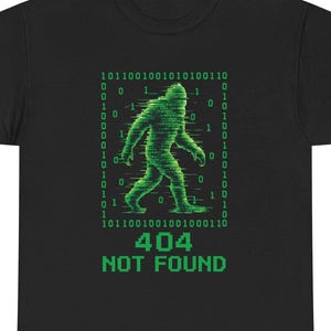 May include: A black t-shirt featuring a green graphic of a Bigfoot silhouette composed of digital glitch effects and binary code. The design is framed by a border of ones and zeros, with the pixelated text 404 NOT FOUND printed below the figure.