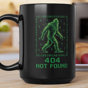 May include: A black ceramic mug featuring a green graphic of a Bigfoot silhouette composed of binary code digits. Below the graphic, the text 404 NOT FOUND is printed in a green pixelated font. The design is centered on the front of the mug.