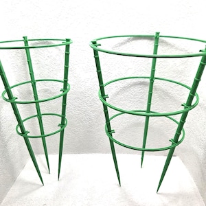 May include: Two green tomato cages. Each cage has a conical shape with three circular support rings and three pointed legs. The cages are made of a durable, weather-resistant material, perfect for supporting growing plants in a garden.