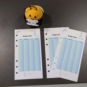 Könnte beinhalten: Eine gehäkelte gelb-schwarze Bienenfigur, die ein kleines Schild mit der Aufschrift Bee Happy hält, dazu drei weiße Papier-Budgetblätter mit blauem Verlaufsraster, Meerjungfrauen-Illustrationen und dem Text Budget Sheet.