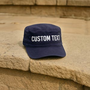 May include: A navy blue military-style cap with the words "CUSTOM TEXT" in white block letters across the front. The cap is sitting on a stone surface. This hat is a customizable accessory.