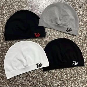 Op de afbeelding: Vier mutsen in zwart, wit en grijs. Elke muts heeft het woord "Tribe" in een contrasterende kleur op de voorkant geborduurd. De mutsen zijn gemaakt van een zacht, rekbaar materiaal.