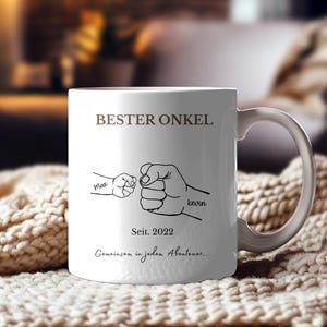 Può includere: Tazza in ceramica bianca con la scritta "BESTER ONKEL" e un disegno di due pugni che si toccano. La tazza include anche i testi "Max", "Kevin", "Seit. 2022" e "Gemeinsam in jedem Abenteuer..."
