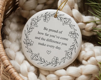 May include: A white ceramic ornament with a floral wreath design and the text "Be proud of how far you've come and the difference you make every day." The ornament hangs from a gold string and is set in a woven basket.